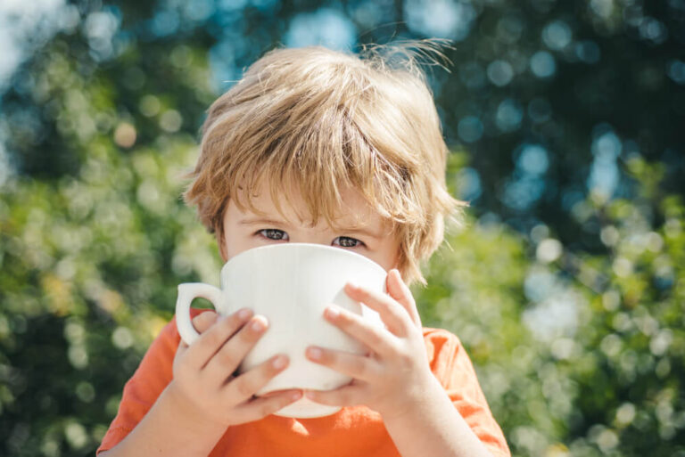 A little toddler is sitting in a garden and is drinking from coffee cup