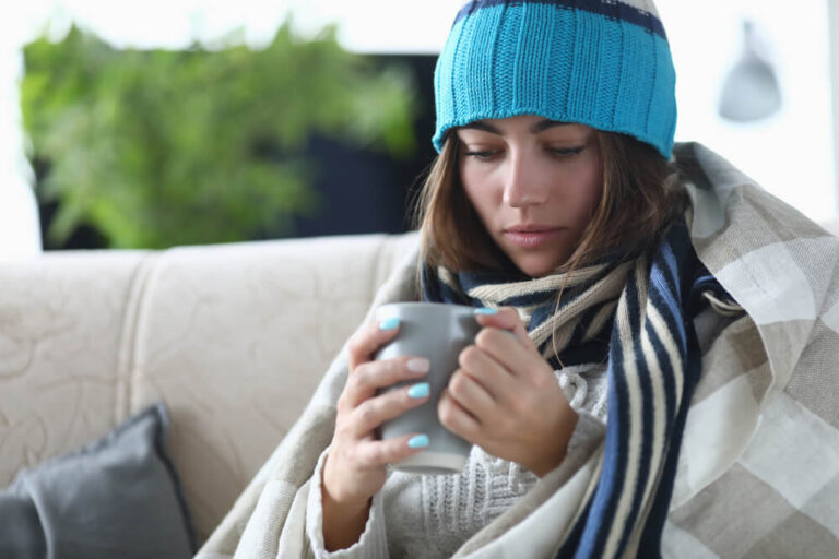 Young girl with flu in hat and scarf under covers holding coffee