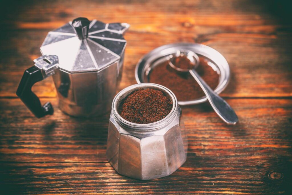Moka pot with ground coffee inside and with spoon beside it.