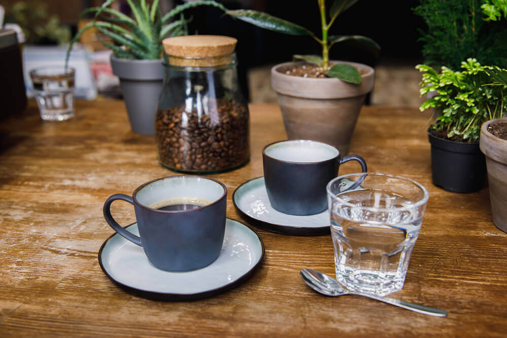 a cup of coffee and a cup of water in a table with spoon and coffee beans