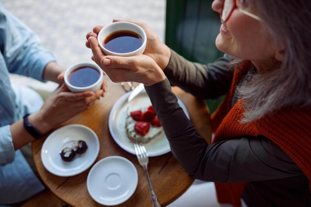 Smiling mature woman with best friend hold cups of hot tea sitting at small table in cozy cafe