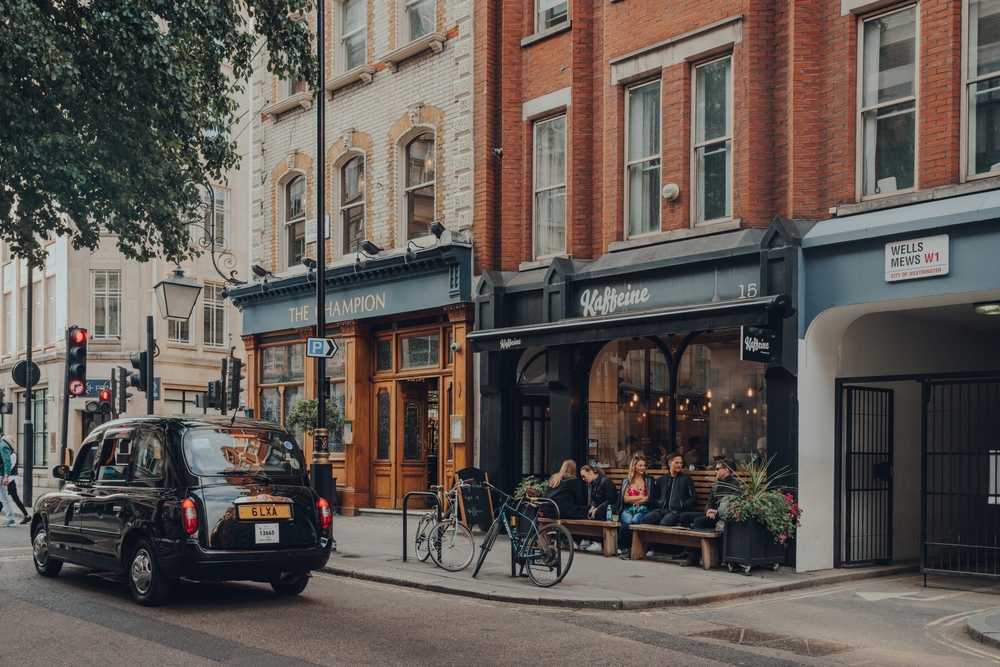 Kaffeine coffee shop in Fitzrovia, diverse area of Central London dotted with restaurants, hotels, galleries and old-school pubs, people outside, taxi drives past.