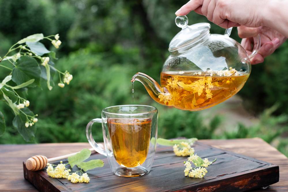 Female hands pouring linden tea from a glass teapot into a cup in the garden, healthy herbal tea, close up.