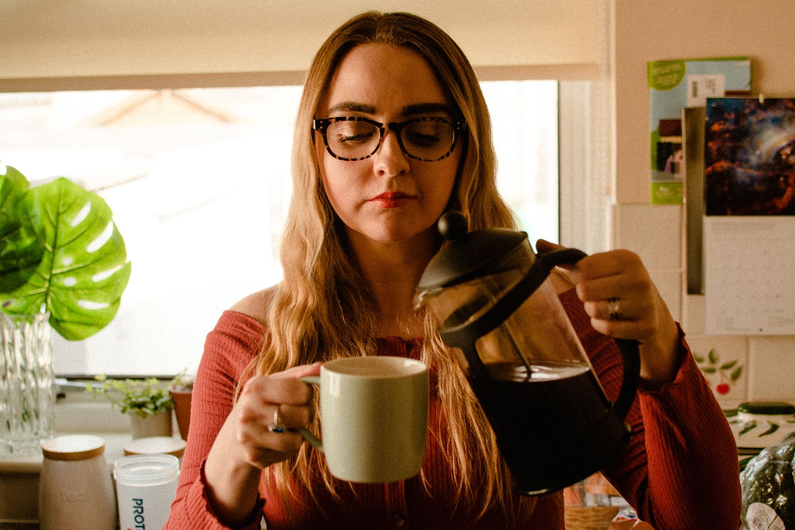 A woman pours coffee from a French press into a mug in a bright kitchen.