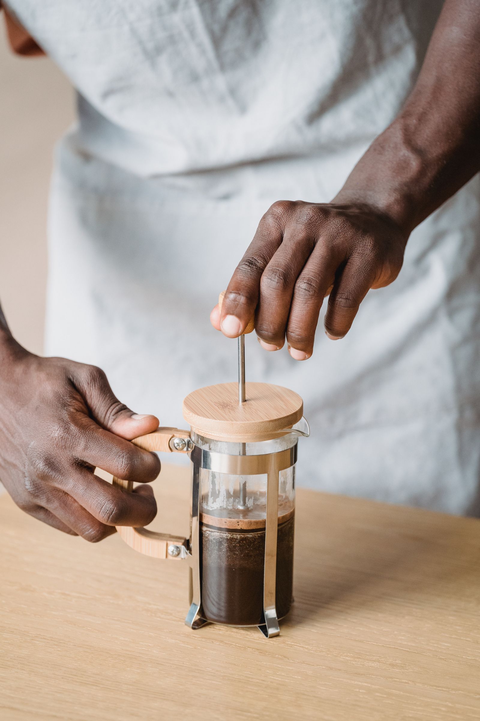 Hands press the plunger of a generic French press on a wooden table.